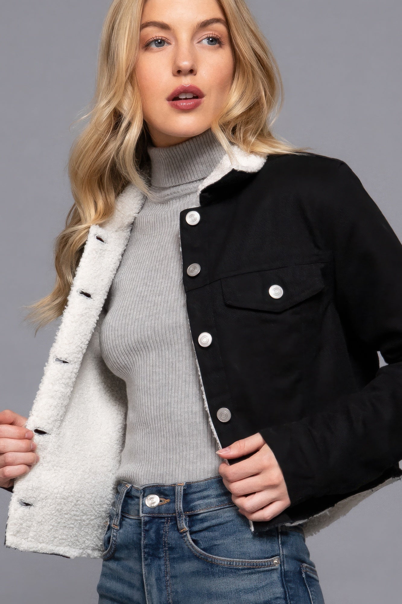 Woman wearing a black jacket with white lining, gray turtleneck, and blue jeans on a gray background